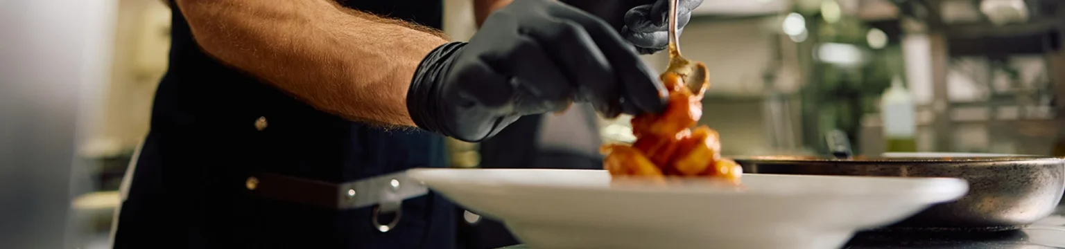 Chef plating a dish coated in a glossy sweet and spicy sauce in a commercial kitchen.