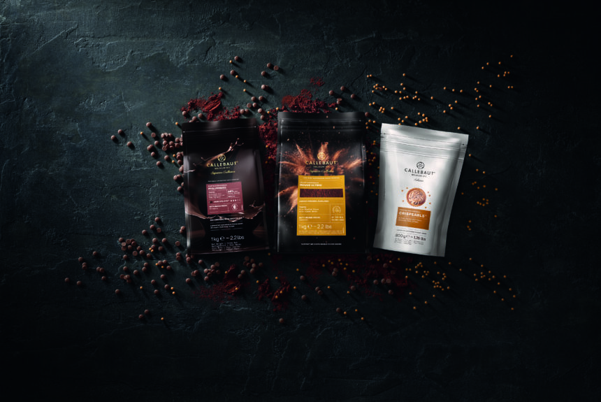 Three Callebaut products displayed with chocolate callets, cocoa powder and Crispearls on a dark textured background.