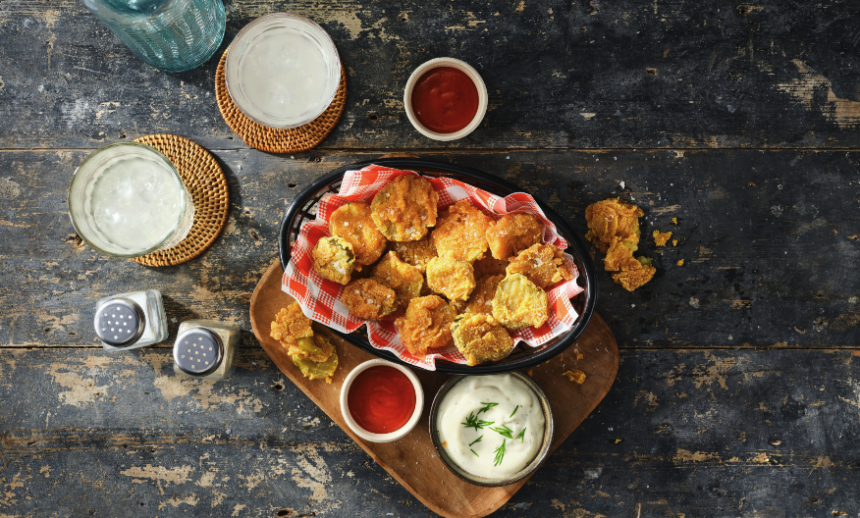 Crispy fried pickle chips served with creamy dill dip and red dipping sauce on a rustic bar table.