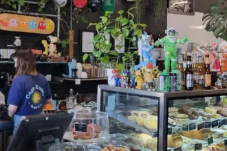 A Café Barco barista preparing coffee behind a counter filled with pastries, plants and colourful decorations.