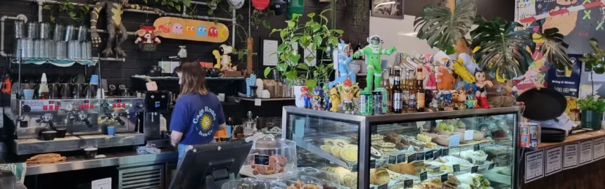 A Café Barco barista preparing coffee behind a counter filled with pastries, plants and colourful decorations.