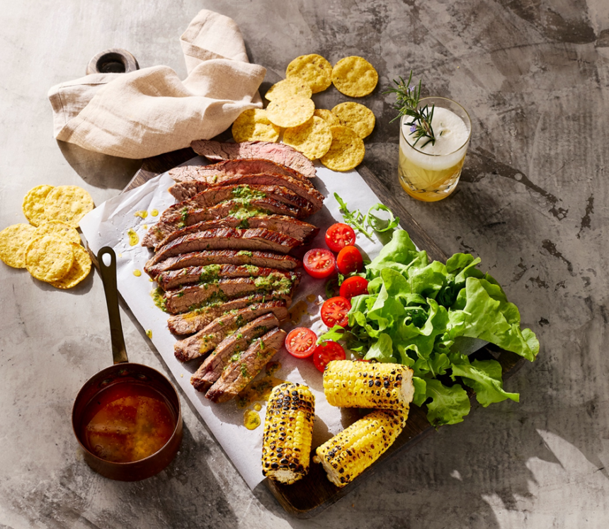 Sliced skirt steak with cowboy butter, grilled corn, lettuce, tomatoes and corn chips on a sharing board.