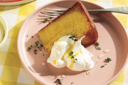 A slice of golden olive oil cake served with whipped cream, fresh herbs and a drizzle of olive oil on a pink plate.