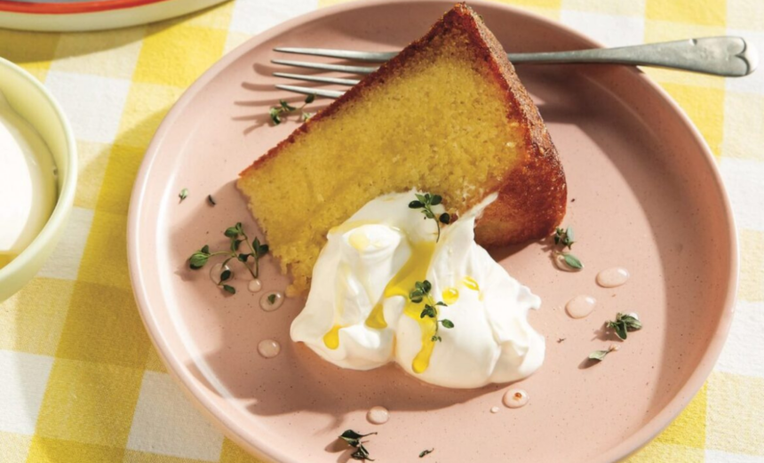 A slice of golden olive oil cake served with whipped cream, fresh herbs and a drizzle of olive oil on a pink plate.