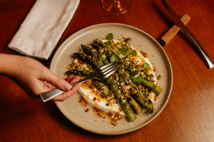 Charred asparagus served over whipped feta with hot honey, seed crumb and fresh herbs as a diner reaches in with a fork.