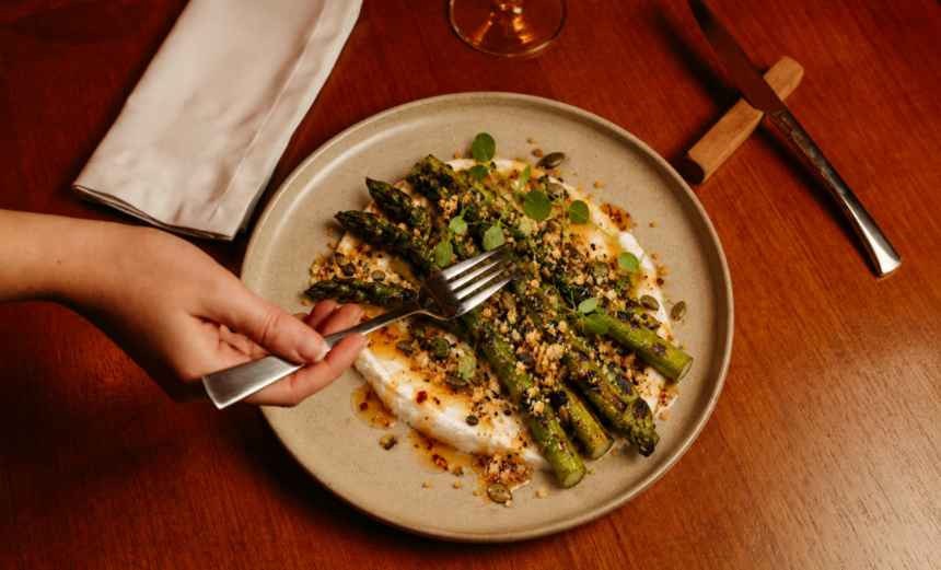 Charred asparagus served over whipped feta with hot honey, seed crumb and fresh herbs as a diner reaches in with a fork.