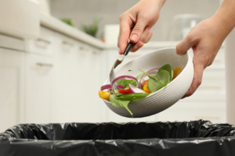 Person scraping leftover salad from a bowl into a lined rubbish bin.