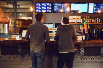 Two customers standing at a quick-service restaurant counter beneath digital menu boards.