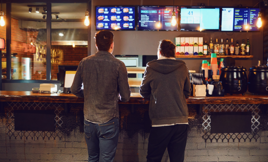 Two customers standing at a quick-service restaurant counter beneath digital menu boards.