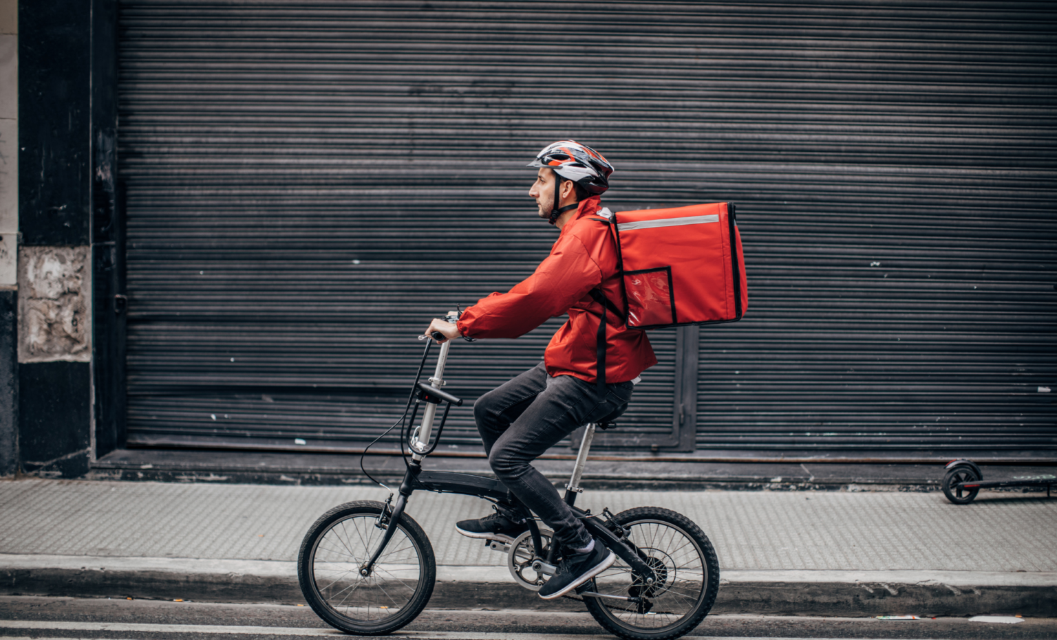 Food delivery rider wearing a helmet and carrying a large red insulated bag while cycling along a street.