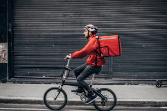 Food delivery rider wearing a helmet and carrying a large red insulated bag while cycling along a street.