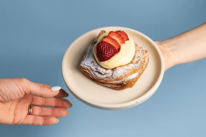A Strawberry Cheesecake Danish dusted with icing sugar and topped with cream and fresh strawberries, served on a plate against a blue background.