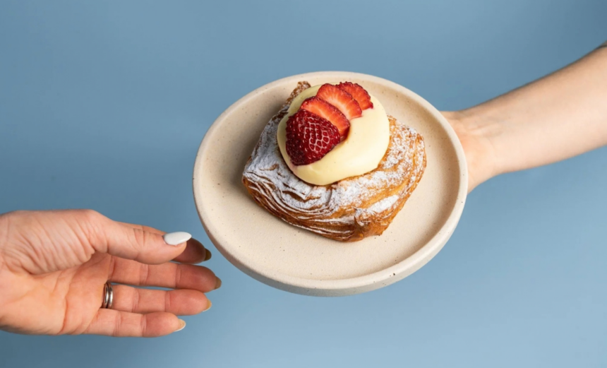 A Strawberry Cheesecake Danish dusted with icing sugar and topped with cream and fresh strawberries, served on a plate against a blue background.