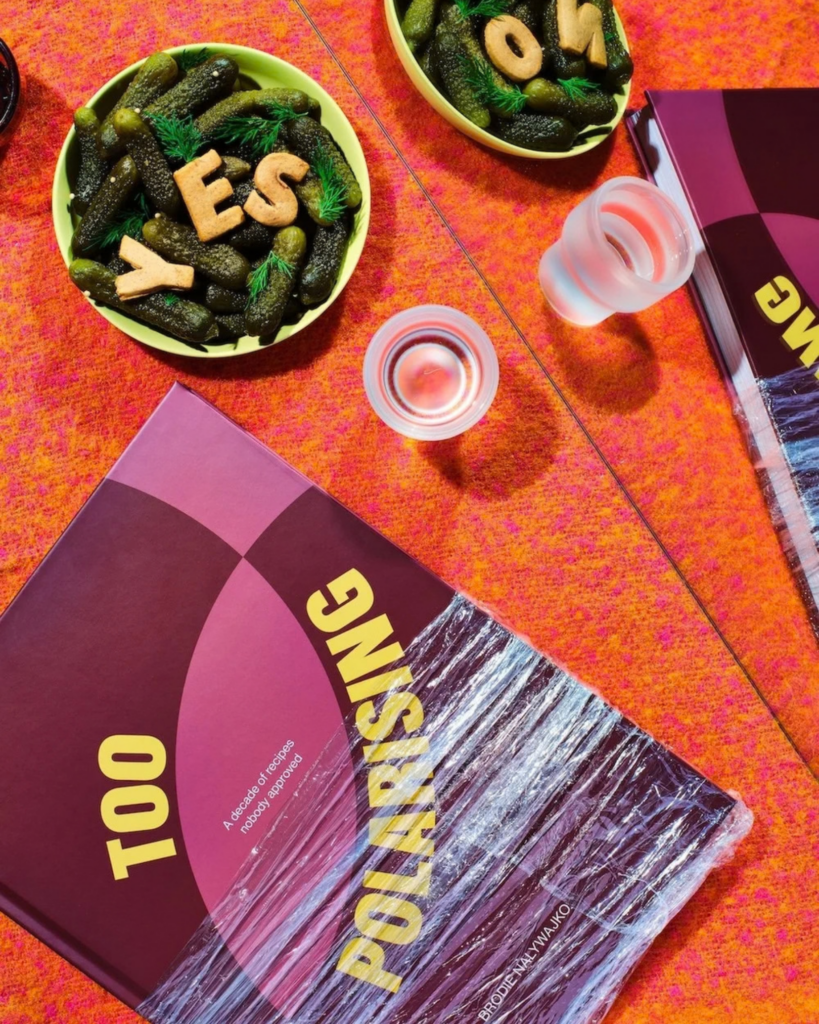 Copies of Too Polarising by Brodie Nalywajko displayed on a bright orange table alongside bowls of pickles and drinks.