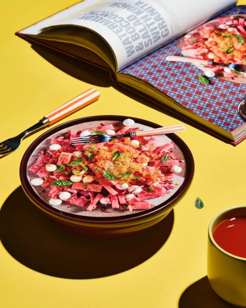 Colourful dish styled on a bright yellow table beside an open copy of Brodie Nalywajko’s cookbook Too Polarising.