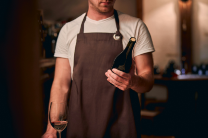 Restaurant staff member in an apron holding a bottle of red wine beside a partially filled wine glass.