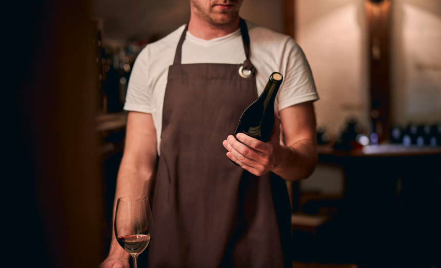Restaurant staff member in an apron holding a bottle of red wine beside a partially filled wine glass.